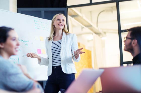 Anaplan_Web_2020_SolutionsPages_Marketing_FINAL Woman presenting to coworkers in front of dry erase board